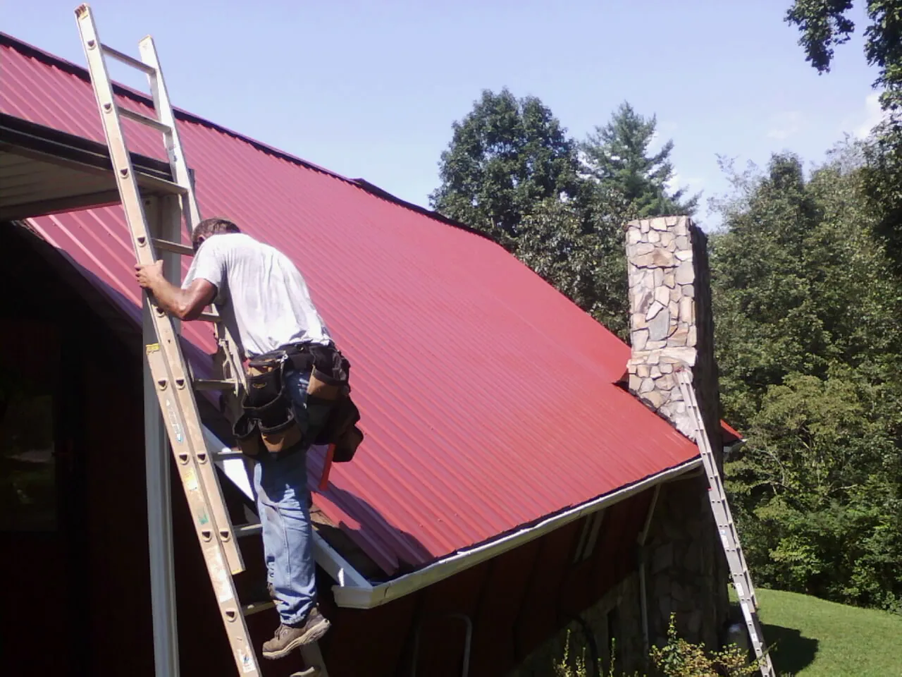 Roofer on a ladder beside a red metal roof and stone chimney — Soffit & Fascia Repair by Roofing Brothers in Peachland